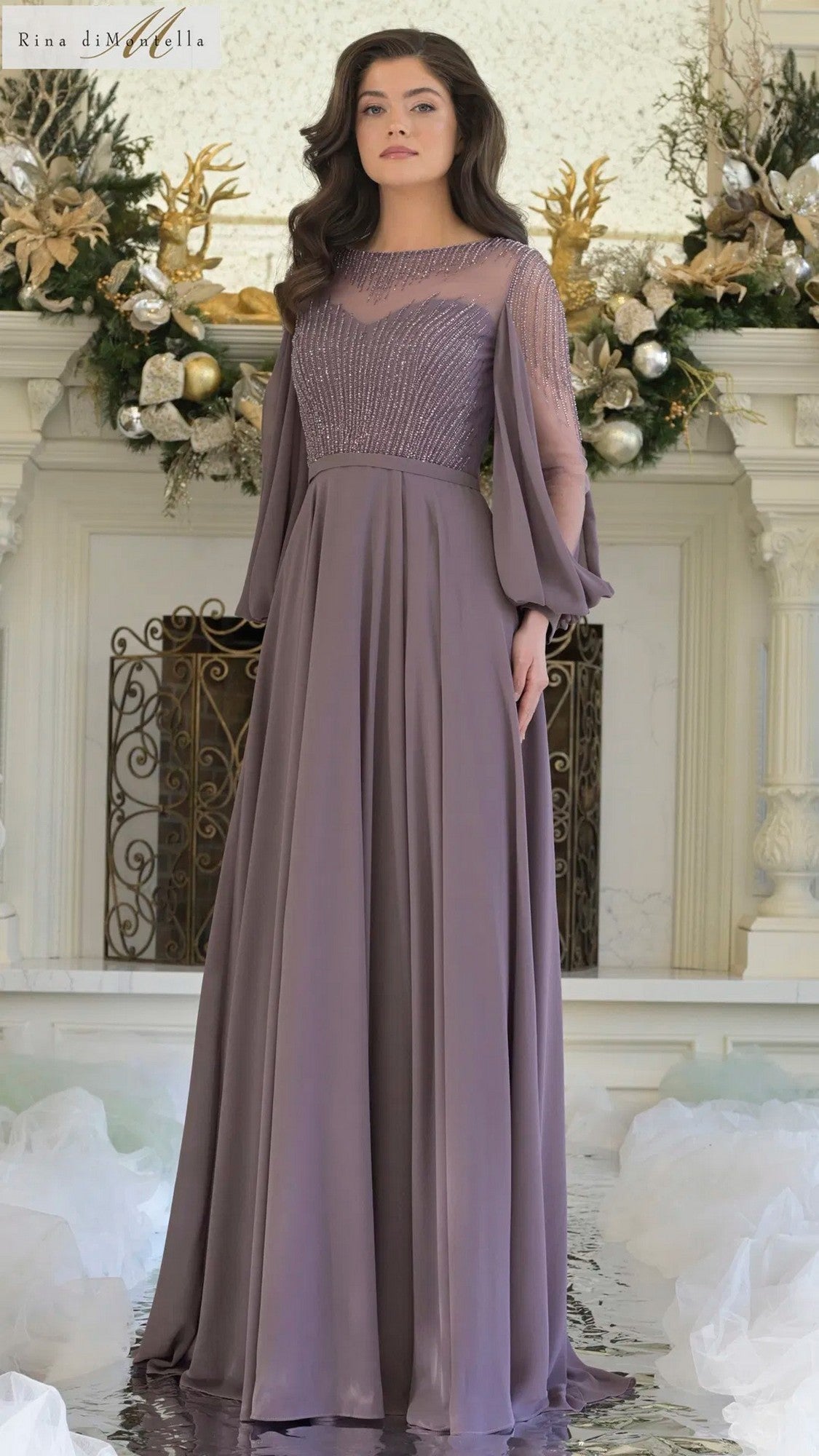 A woman stands indoors by a decorated fireplace wearing the Rina Di Montella RD3374 long formal dress in Victorian Lilac, featuring sheer sleeves and a beaded bodice.